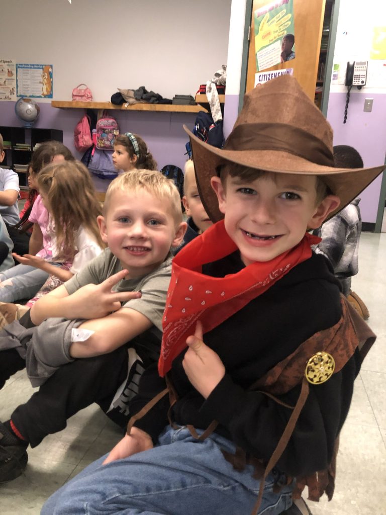 Smiling students sitting on the classroom floor, one dressed in a cowboy hat and bandana